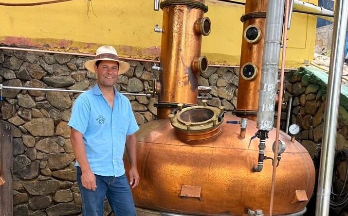 Homem vestido de camisa azul, calça jeans e chapéu. Ao seu lado, há um caldeirão para decantar a cana-de-açúcar