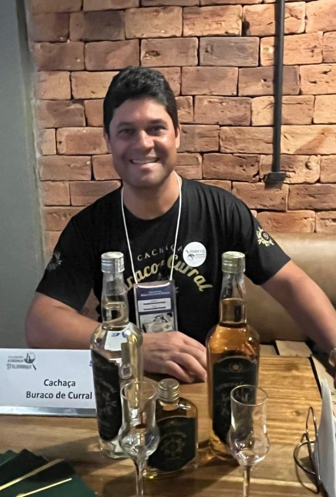 Homem com camiseta preta, sentado e à sua frente, garrafas de cachaça que ele produz