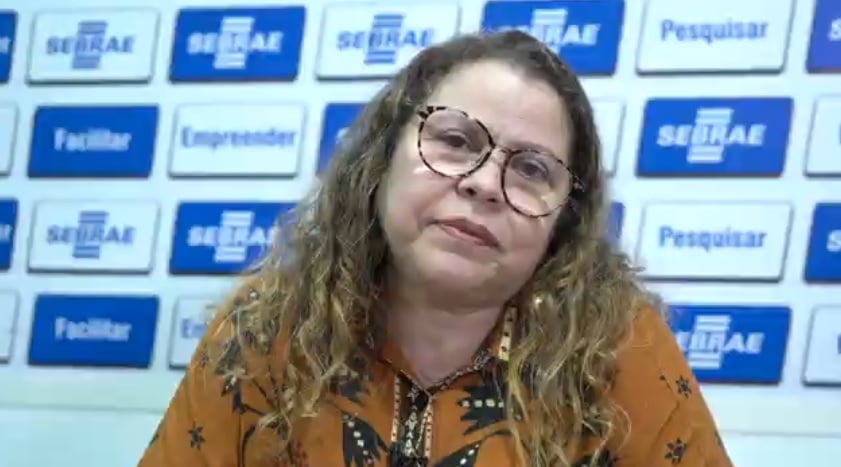 Na foto, há uma mulher que usa óculos e está com uma blusa na cor ferrugem. 