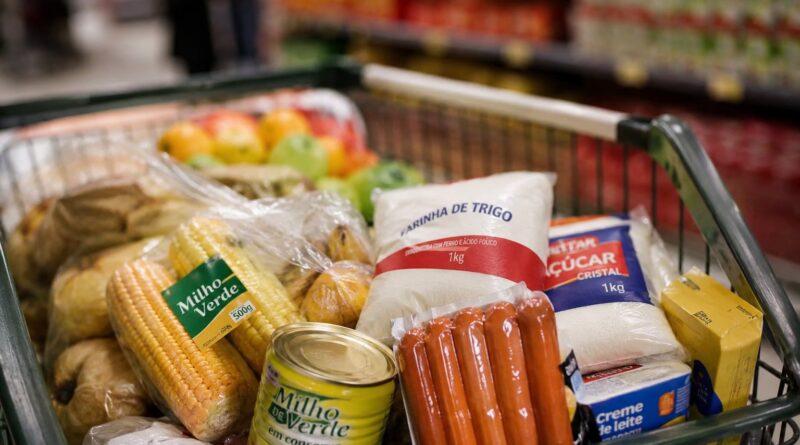 Imagem mostra carrinho de supermercado com comidas típicas de festa junina, como milho, salsicha e açúcar
